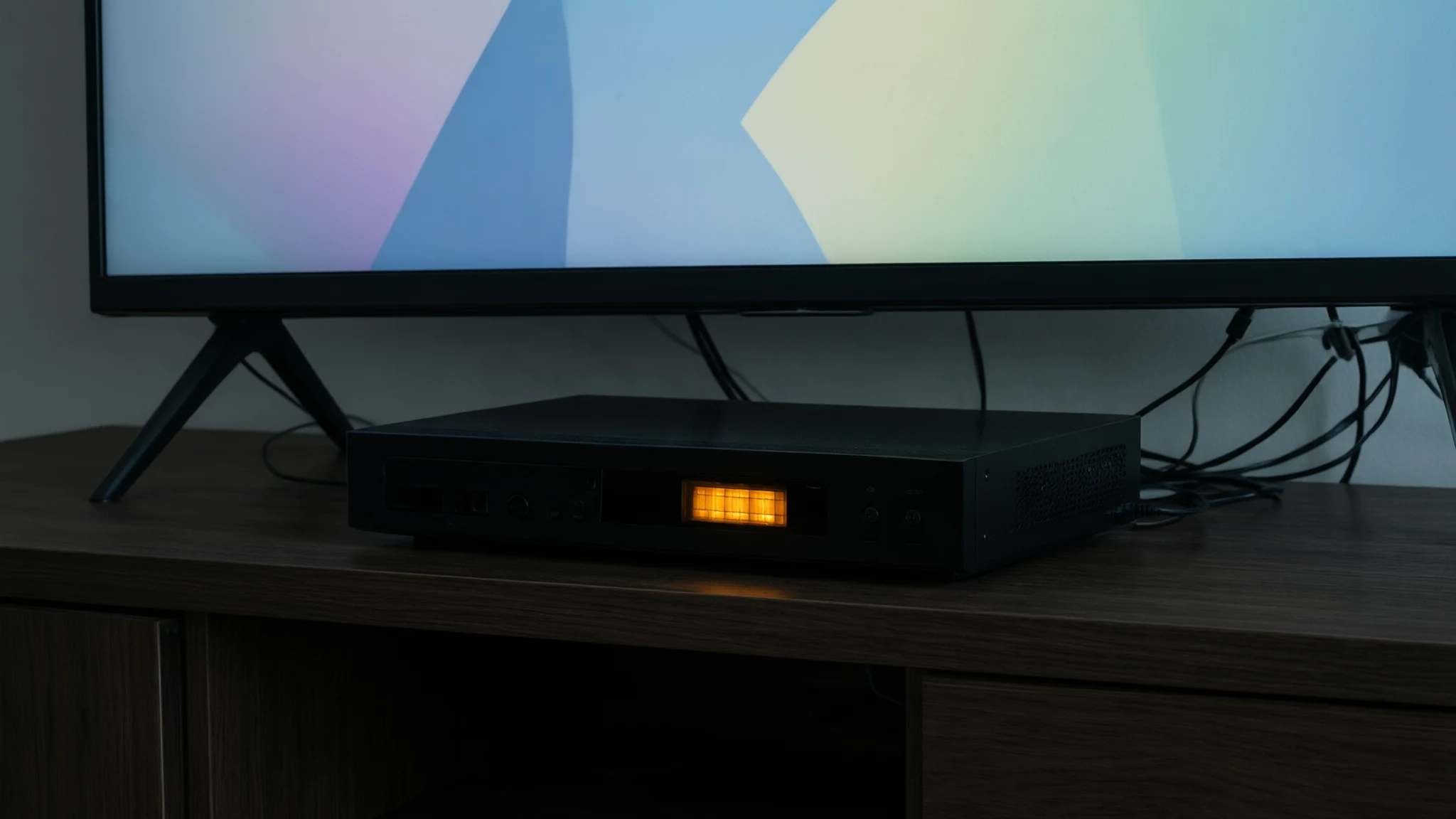 Slim black set-top box with a glowing amber display under a TV on a dark wood unit